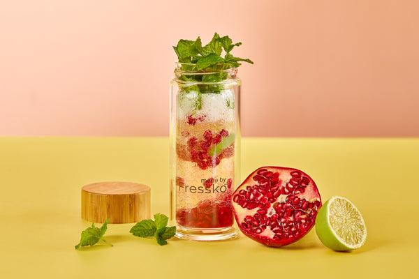 A Glass Fressko Flask is filled with ice, pomegranate seeds, mint and a slice of lime. There is half a pomegranate, half a lime and some mint leaves infront of the flask. The background is peach and yellow.