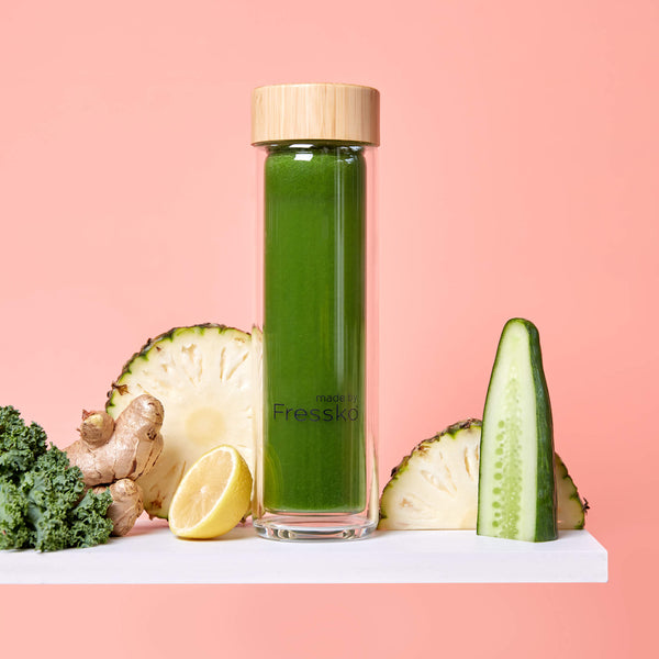 Green juice in Fressko glass flask surrounded by cucumber, pineapple and kale
